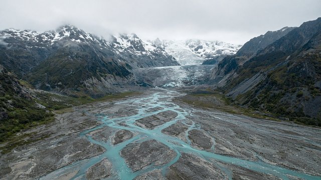 Aerial view of a turquoise braided glacial river weaving across a wide grey gravel valley between dark mountains, glacier tongue in the distance, soft overcast light, original AI nature wallpaper